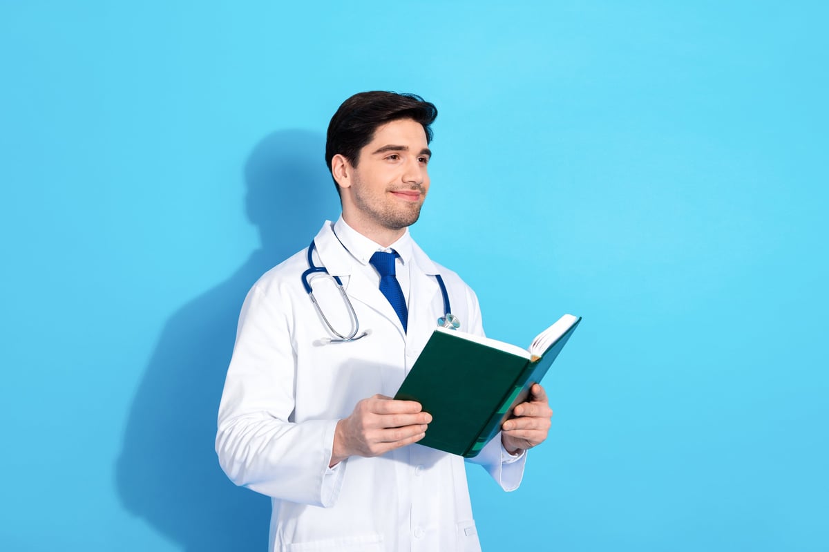 A male doctor in a white coat and stethoscope against a blue background reading a book, symbolizing healthcare education and professionalism