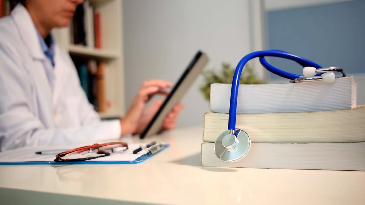 Doctor in a white coat checking medical information on a digital tablet, with a stethoscope on a stack of books